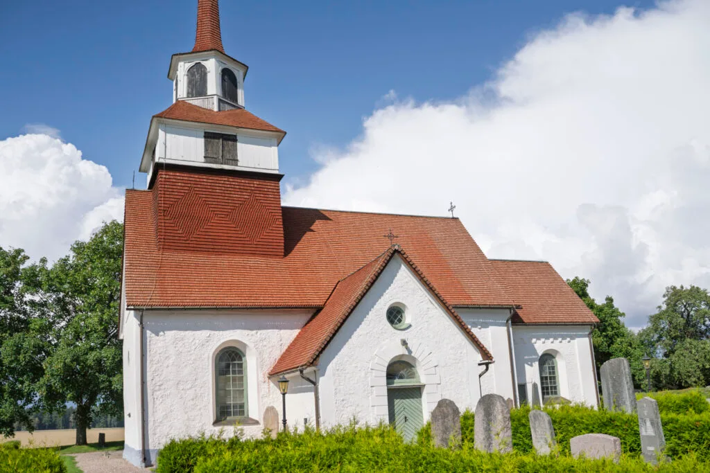Järstads kyrka med vitputsade väggar, rött tegeltak och klockstapel, omgiven av kyrkogård med gravstenar och grönska.