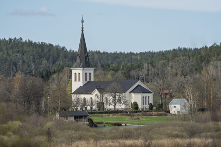 Hannäs kyrka i Åtvidabergs kommun, en vit kyrka från 1885 med spetsigt torn omgiven av grönska och barrskog vid sjö.