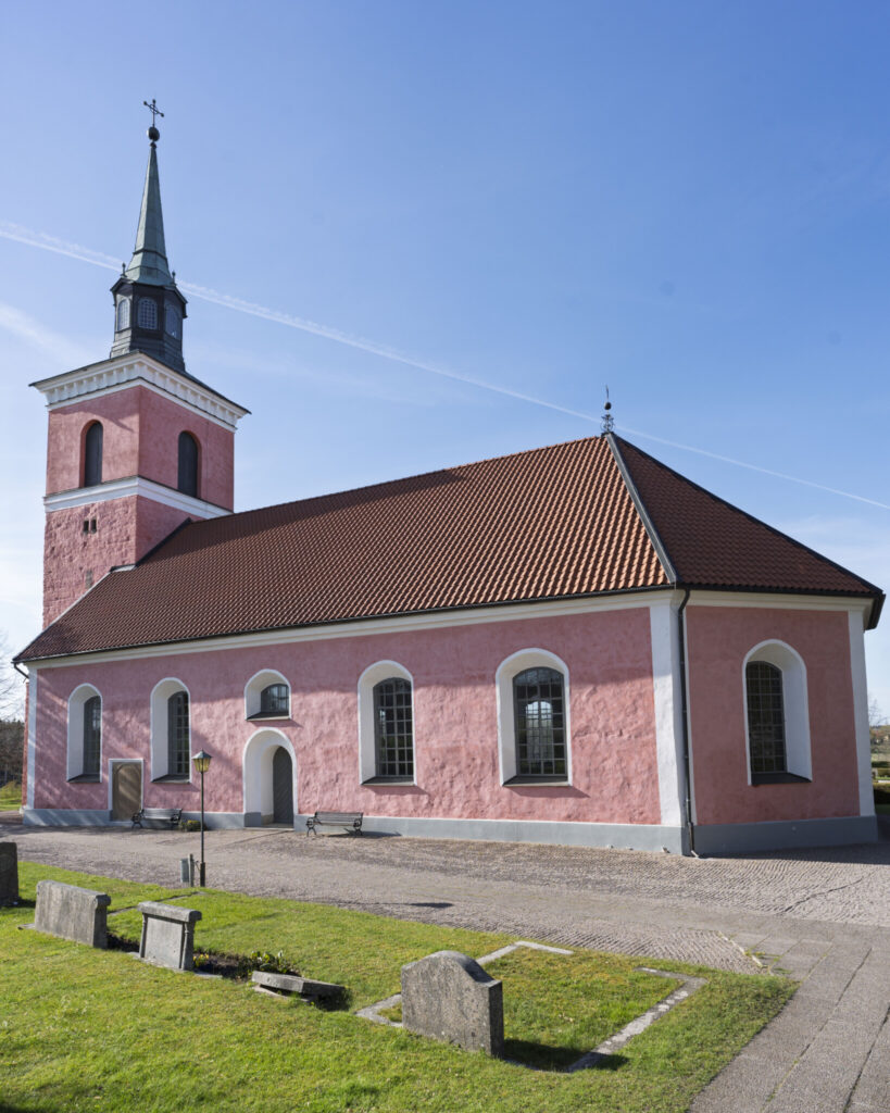Slaka kyrka med sin karaktäristiska rosaputsade fasad, brunt tegeltak och kyrktorn mot klarblå himmel, omgiven av kyrkogård.