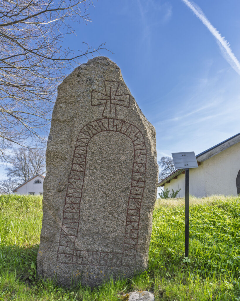 Runsten med ristade runor och kors på gräskulle vid Slaka kyrka i Östergötland, mot blå himmel och informationsskylt