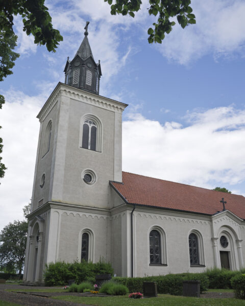 Vitputsad Ledbergs kyrka med torn och spira, omgiven av kyrkogård med gravstenar och grönska, en sommardag i Östergötland