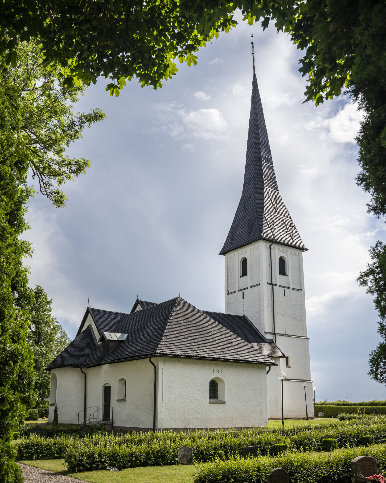 Kaga kyrka med vitputsade väggar, mörkt tak och spetsigt torn omgiven av grön kyrkogård och lummiga träd, Östergötland