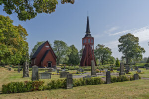 Ulrika kyrka i Östergötland med röd träfasad, fristående klockstapel och kyrkogård omgiven av grönskande lövträd en solig dag