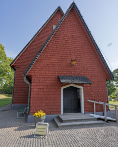 Rödfärgad träkyrka i Ulrika med brant spåntak, öppen dörr och skylt med "Kyrkan är öppen – Välkommen in" samt gula blommor vid entrén.