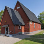 Rödfärgade Ulrika kyrka med brant spåntak och vitputsade fönster, omgiven av kyrkogård och gröna träd en solig sommardag.