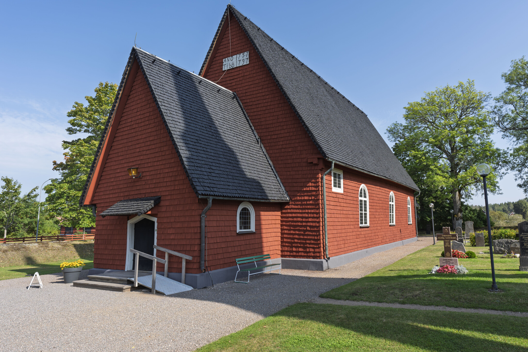 Rödfärgade Ulrika kyrka med brant spåntak och vitputsade fönster, omgiven av kyrkogård och gröna träd en solig sommardag.