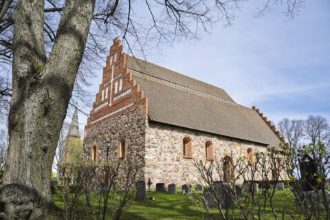 Åtvids gamla kyrka i Åtvidaberg, medeltida stenkyrka med trappgavel i tegel, omgiven av kyrkogård och lövträd en vårdag.