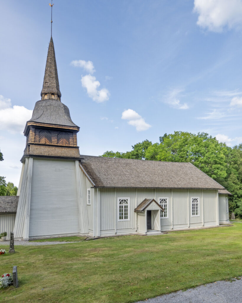 Simonstorps kyrka, en vitpanelad träkyrka från 1600-talet med spåntak och spetsigt klocktorn, omgiven av grön gräsmatta och lövträd.