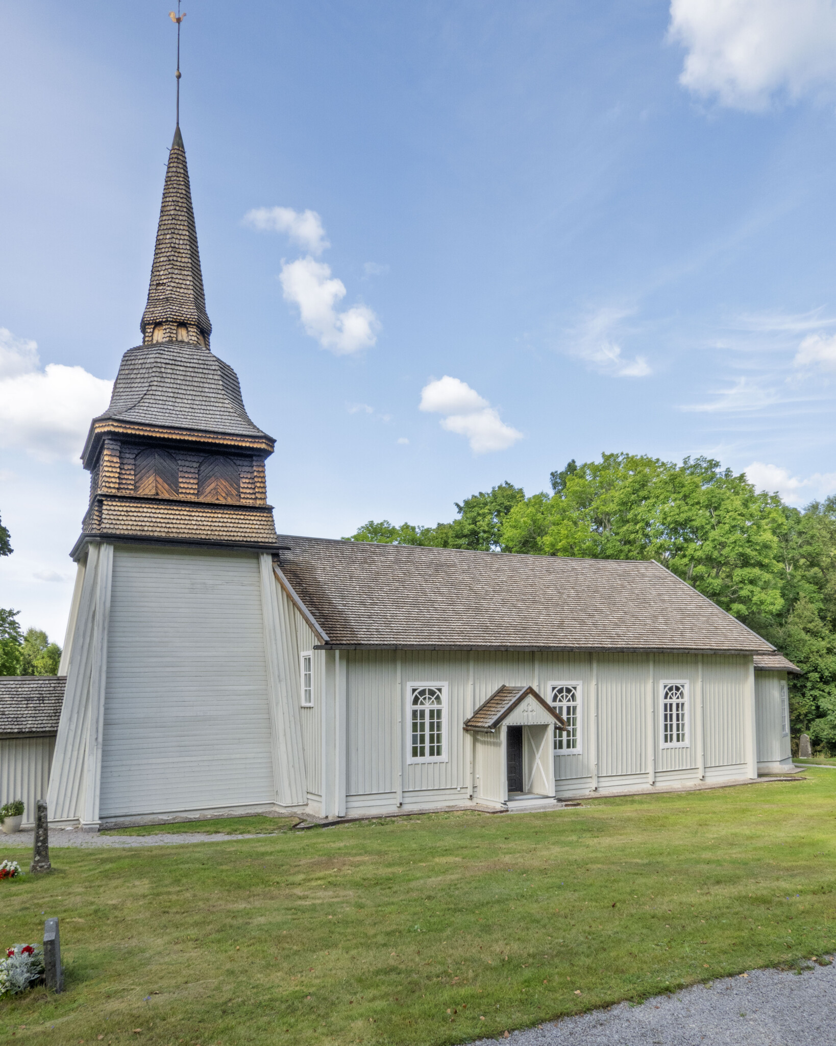 Simonstorps kyrka, en vitpanelad träkyrka från 1600-talet med spåntak och spetsigt klocktorn, omgiven av grön gräsmatta och lövträd.