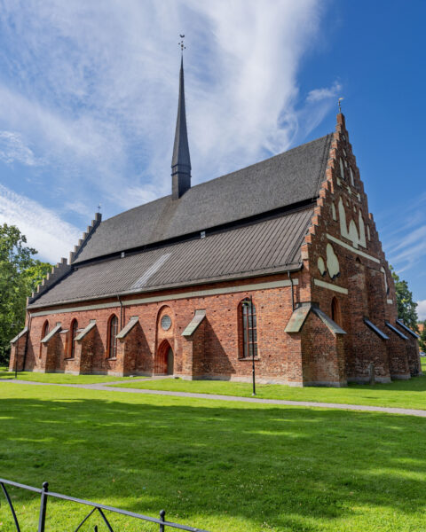 S:t Laurentii kyrka i Söderköping, medeltida tegelkyrka med trappgavel och hög spira omgiven av grön gräsmatta en solig dag