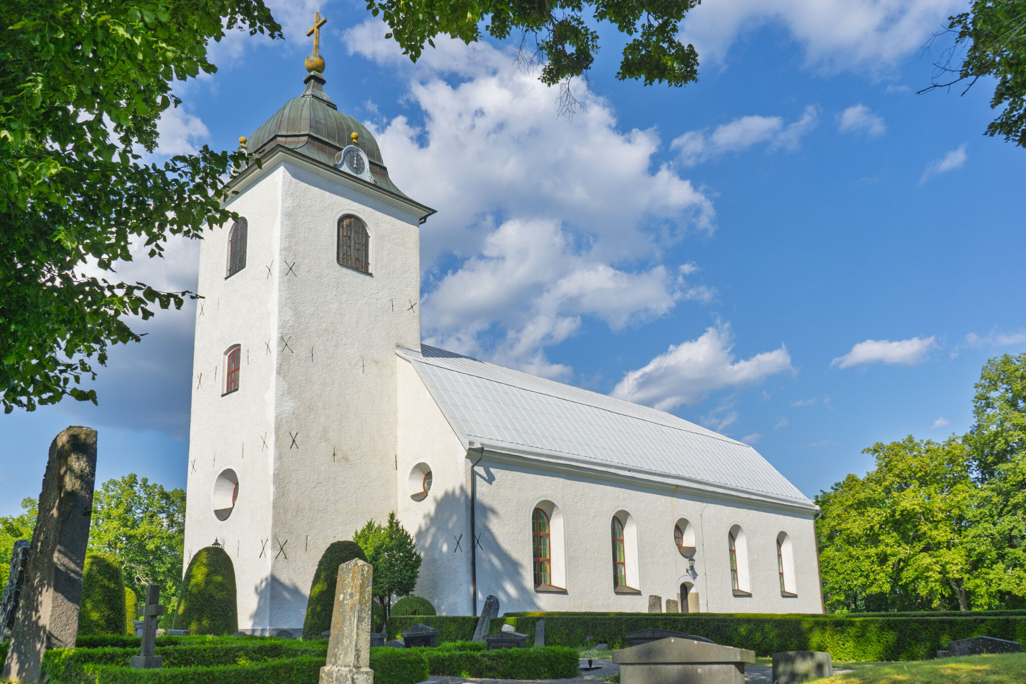 Västra Eneby kyrka med vit putsad fasad, lökformigt torn och kyrkogård omgiven av grönskande träd en solig sommardag.