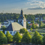 Kisa kyrka med vit fasad och klocktorn omgiven av grönskande träd och kyrkogård, med Kisas småstadsbebyggelse i bakgrunden.