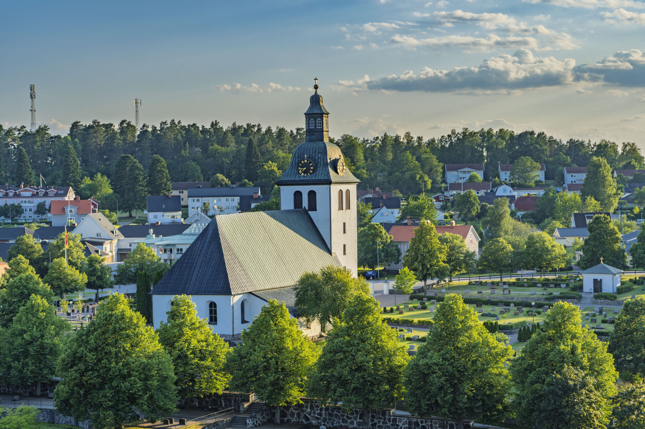 Kisa kyrka med vit fasad och klocktorn omgiven av grönskande träd och kyrkogård, med Kisas småstadsbebyggelse i bakgrunden.