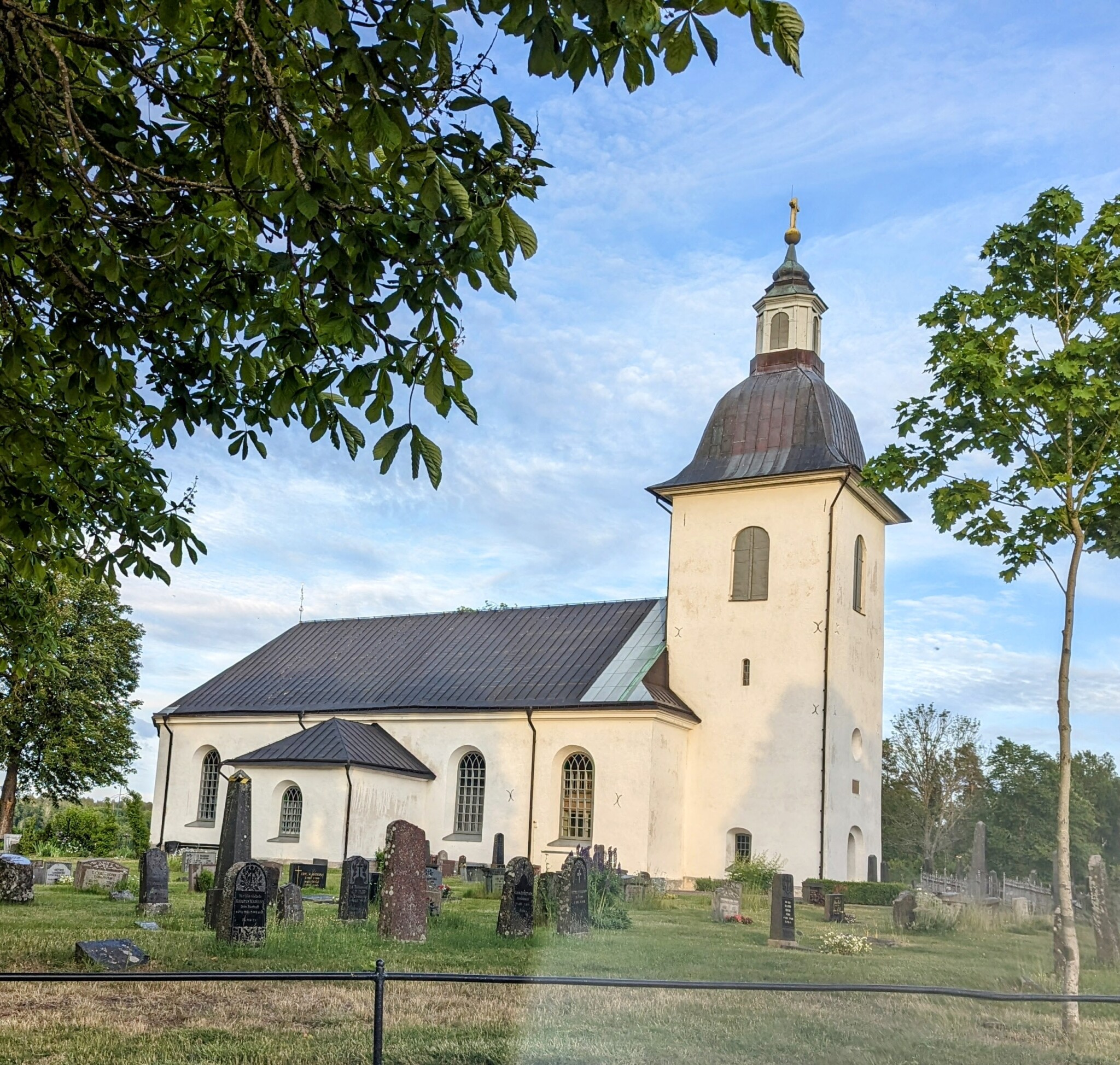 Hycklinge kyrka i Kinda kommun, en vitputsad kyrka från 1792 med kupolformat torntak, omgiven av kyrkogård och grönskande lövträd.