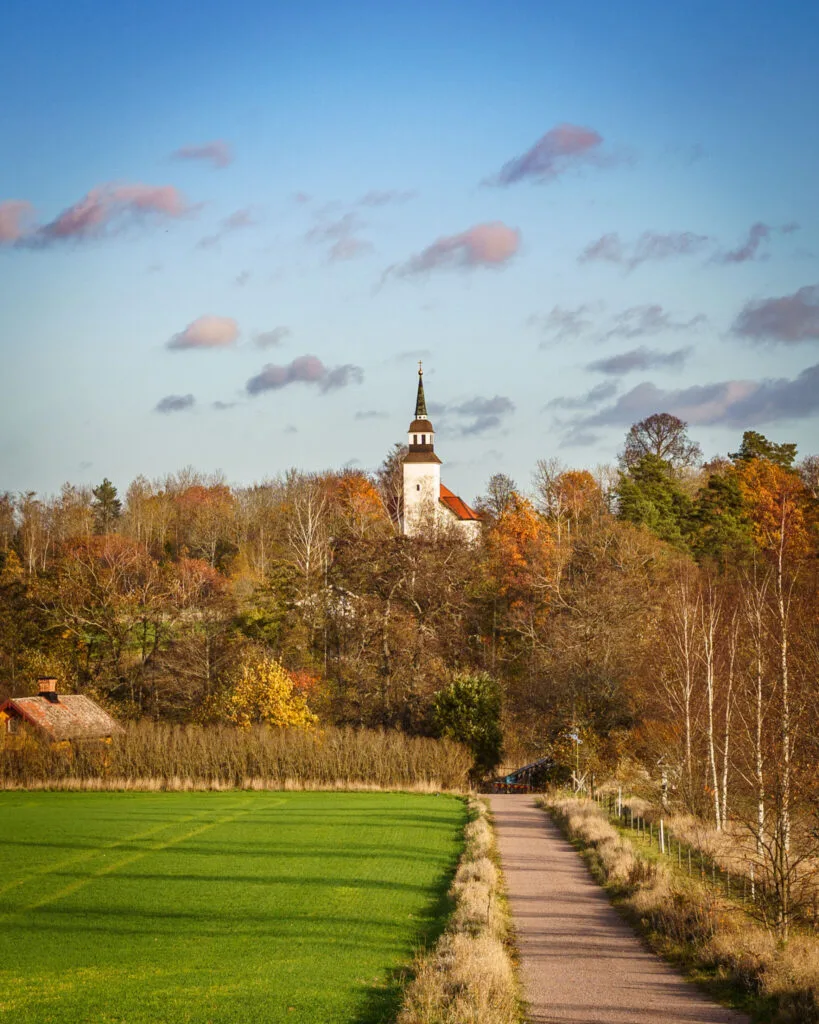 Grusväg längs gröna åkrar i höstlandskap med Landeryds kyrka som reser sig bland färgsprakande lövträd längs hembygdsleden.