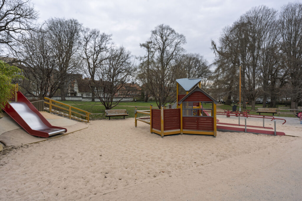 Lekplats i Trädgårdsföreningen i Linköping med rutschkana, lekstuga och sandyta omgiven av lövträd en mulen dag.