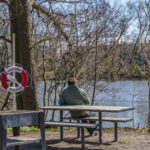 Person sitter på bänkbord vid Stångån på Nykvarnsholmen i Linköping, med livboj och kala träd en tidig vårdag.