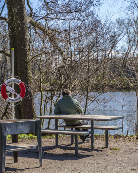 Person sitter på bänkbord vid Stångån på Nykvarnsholmen i Linköping, med livboj och kala träd en tidig vårdag.