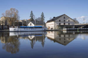 Turistbåt från Rederi AB Kind vid Stångån i centrala Linköping, med Restaurang Stångs Magasin i bakgrunden och spegelblank vattenyta.