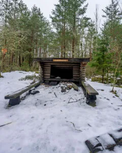 Ullstämmaskogens vindskydd i timmer med eldplats och bänkar omgivet av snötäckt barrskog längs Östgötaleden, Linköping