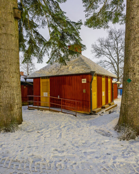 Röd WC-byggnad med gula dörrar vid Dansbanan i Gamla Linköping, omgiven av snötäckt mark och stora träd en vinterdag.