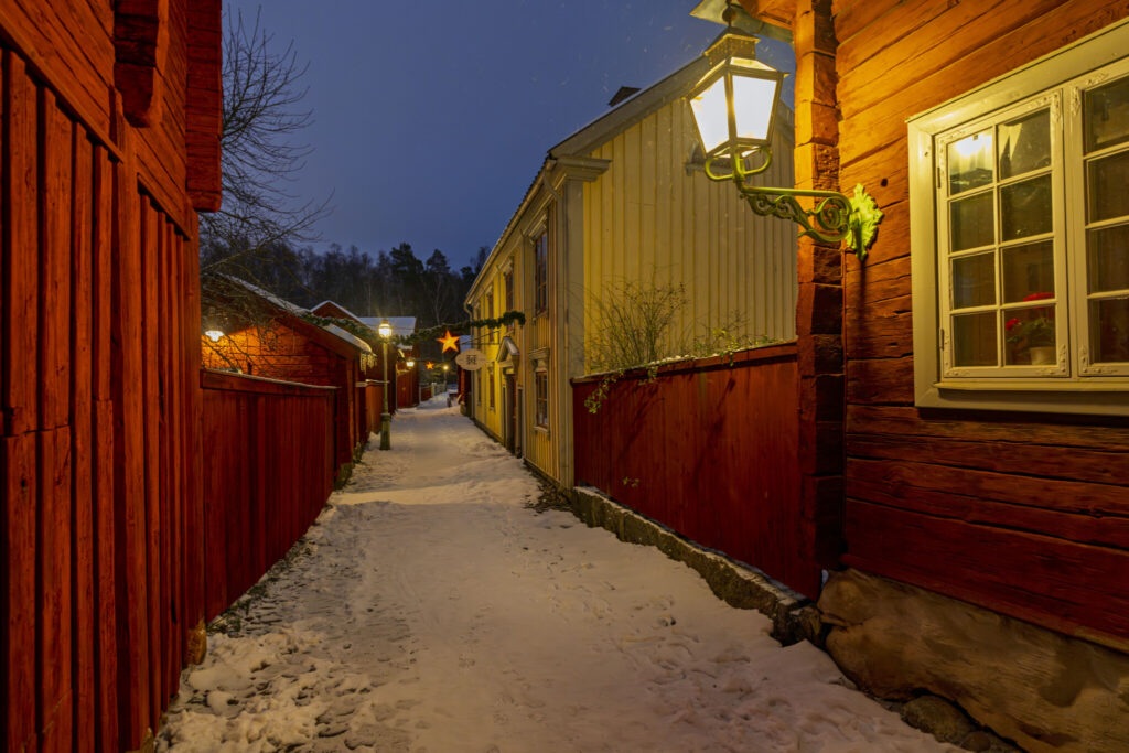 Snötäckt gränd i Gamla Linköping en vinterkvall med faluröda och gula trähus, lyktor och en julstjärna i bakgrunden
