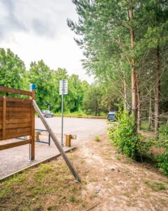 Parkering vid entrén till Motala ströms ravin med trästaket, informationsskylt, parkerade bilar och omgivande tallskog och lövträd.