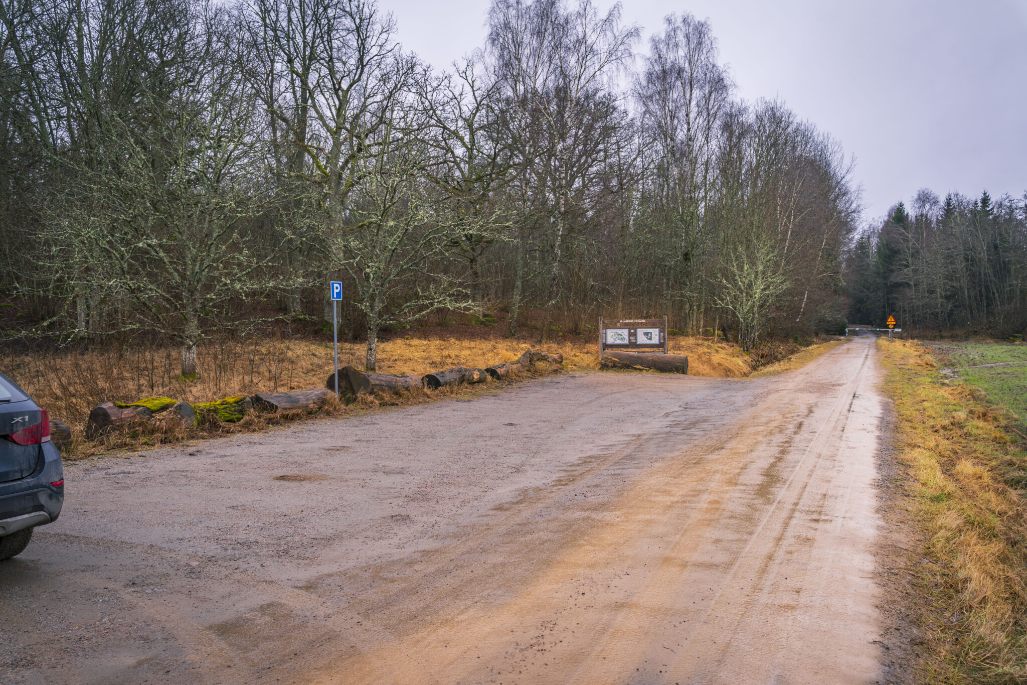 Grusad parkering vid Ullaberg och Mossens våtmarker med parkeringsskylt, informationstavla och stockar som avgränsning, omgiven av lövträd.