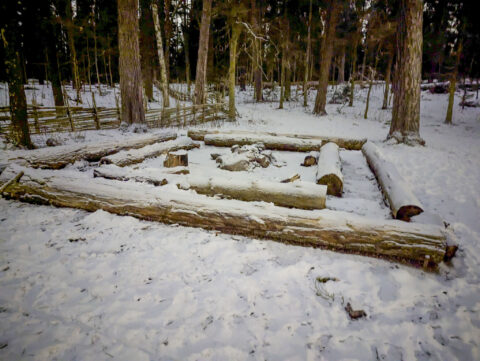 Snötäckt enkel grillplats med stockar som sittplatser i Vallaskogen, Linköping, omgiven av vinterskog och gärdsgård.