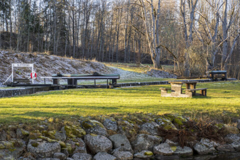 Slattefors sluss vid Kinda kanal en tidig vårmorgon med frostigt gräs, slussluckor, skylt och rastplats med bänkbord omgivna av lövträd.