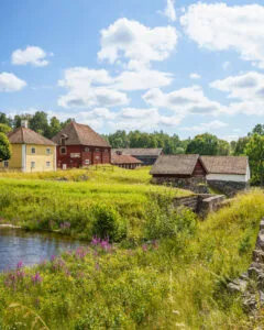 Idyllisk sommarvy över Godegård med gult och rött bruk, stenmurar och ekonomibyggnader vid ett vattendrag omgivet av grönska och lila blommor.