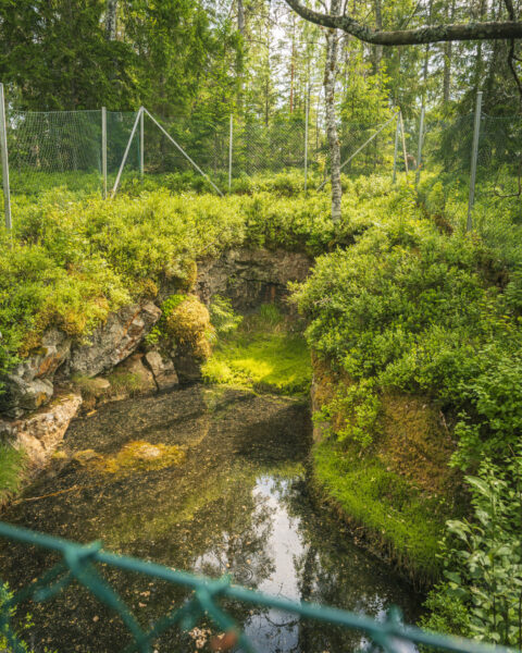Vattenfylld gruvöppning vid Rådsla gruva omgiven av frodig grönska och mossklädda klippor, inhägnad med stängsel längs Rådslaleden