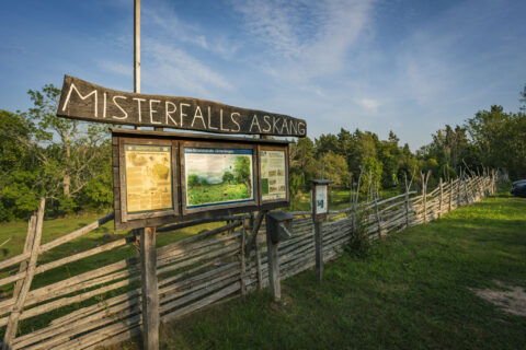 Skylt och informationstavlor vid Misterfalls Askäng i Kinda kommun, omgivna av grönt gräs, trägärdsgård och lummig grönska.