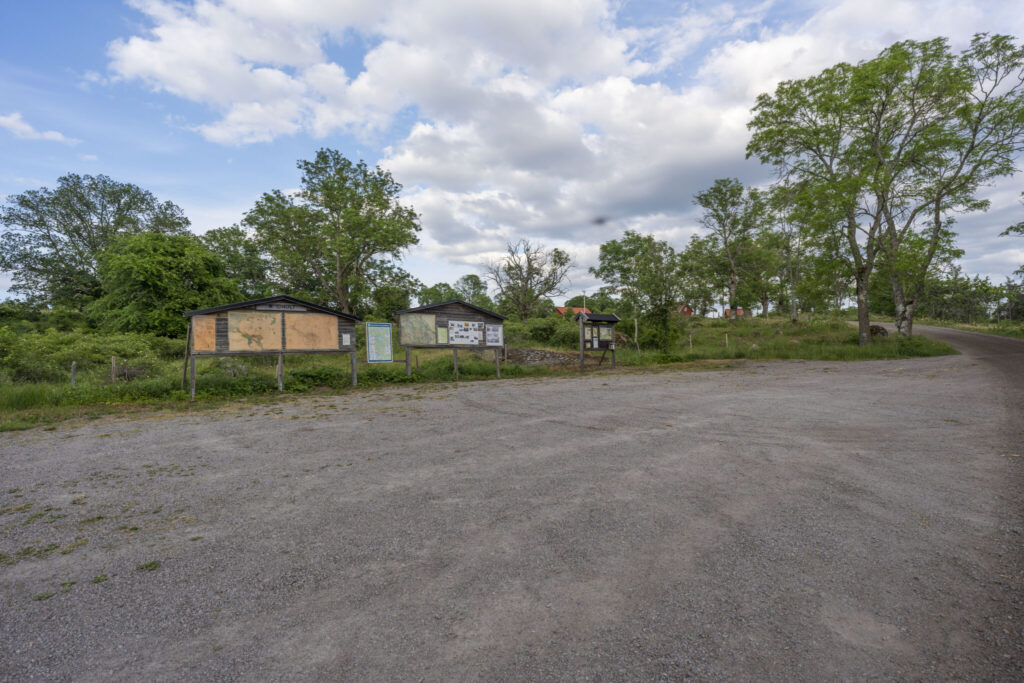 Grusad parkering vid Idhult naturreservat i Kinda kommun med informationstavlor, en liten byggnad och lövträd i sommargrönska.