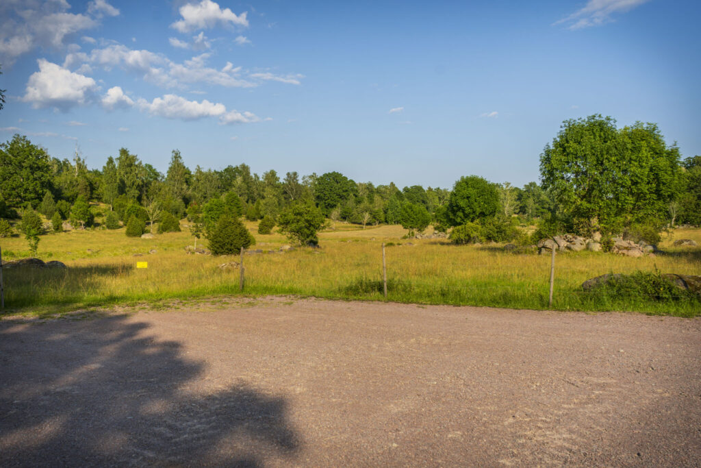 Grusad parkeringsplats vid Föllingsö med utsikt över det öppna odlingslandskapet, gräsmarker och lövträd en sommardag.