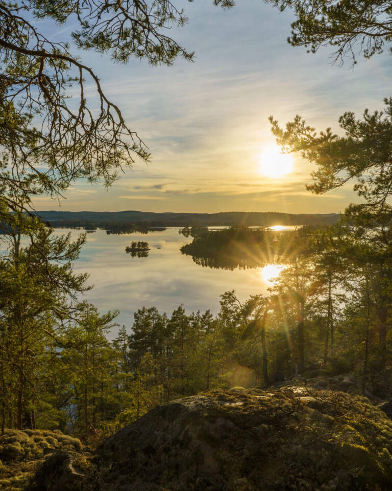 Solnedgång över sjön Åsunden sedd från en klippavsats i Boda naturreservat, med tallar och skogsklädda öar i stilla vatten.