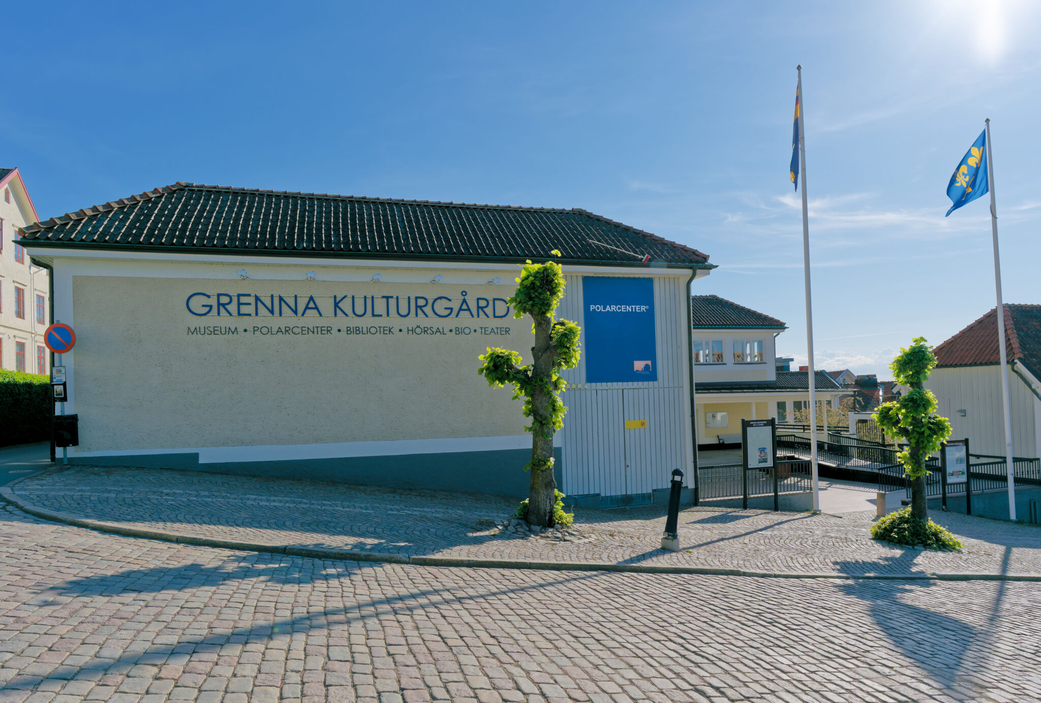 Grenna Kulturgård med museet och Polarcenter i soligt väder, omgiven av kullerstensbelagd torgyta och flaggstänger i Grenna.