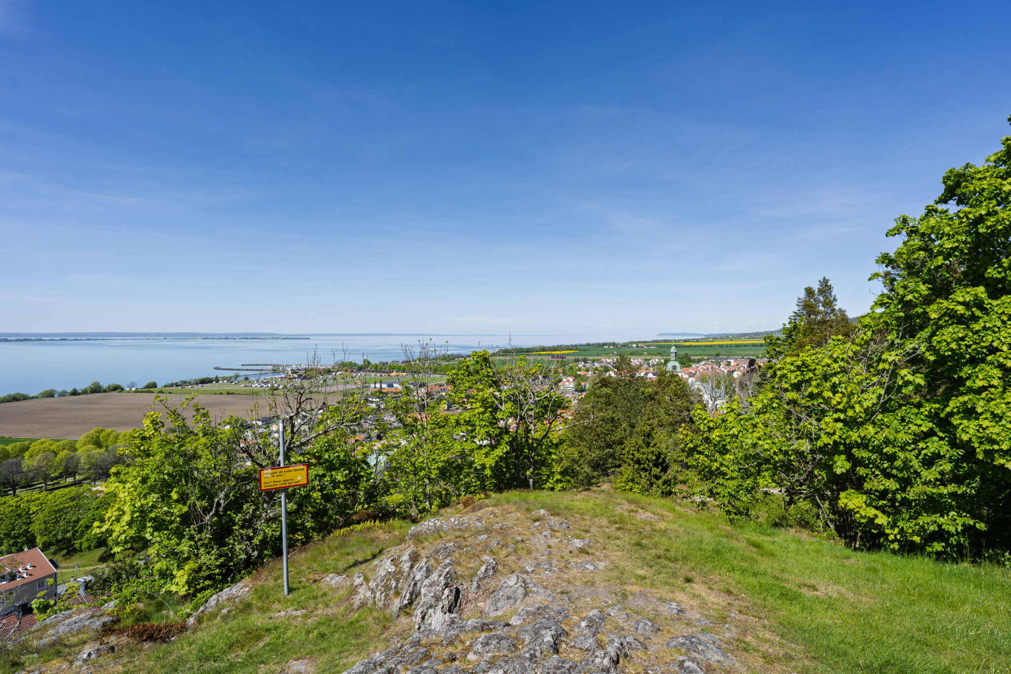 Utsikt från Björkmanskulle över Gränna stad och Vättern en solig sommardag, med grönskande träd i förgrunden.