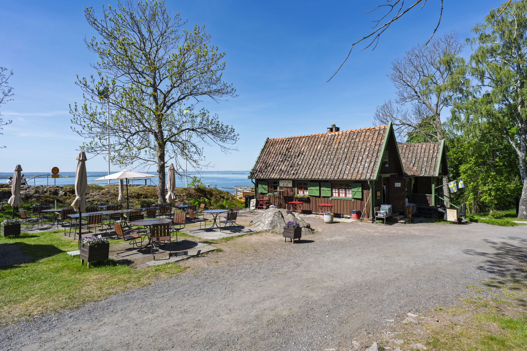 Grännabergets Kaffestuga, en mörk timmerbyggnad med uteservering vid havet, omgiven av grönska en solig sommardag.