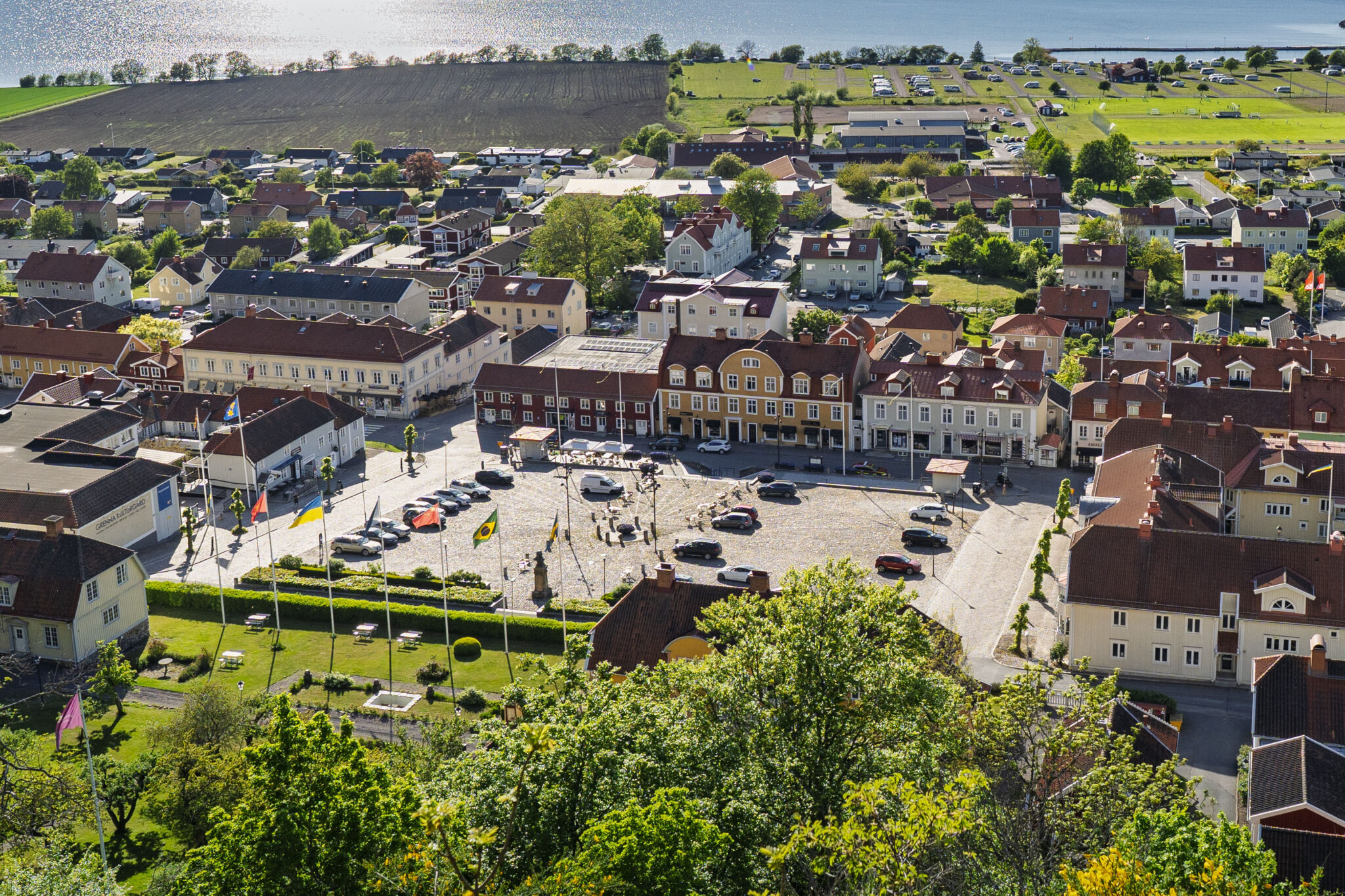 Vy över Gränna torg med omgivande byggnader, flaggstänger och sjön Vättern i bakgrunden.