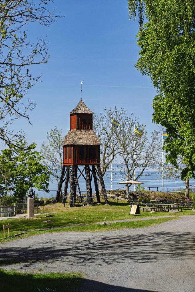 Röd träklocktorn på pålar vid Grännabergets friluftsmuseum, omgiven av gröna träd med Vättern i bakgrunden.