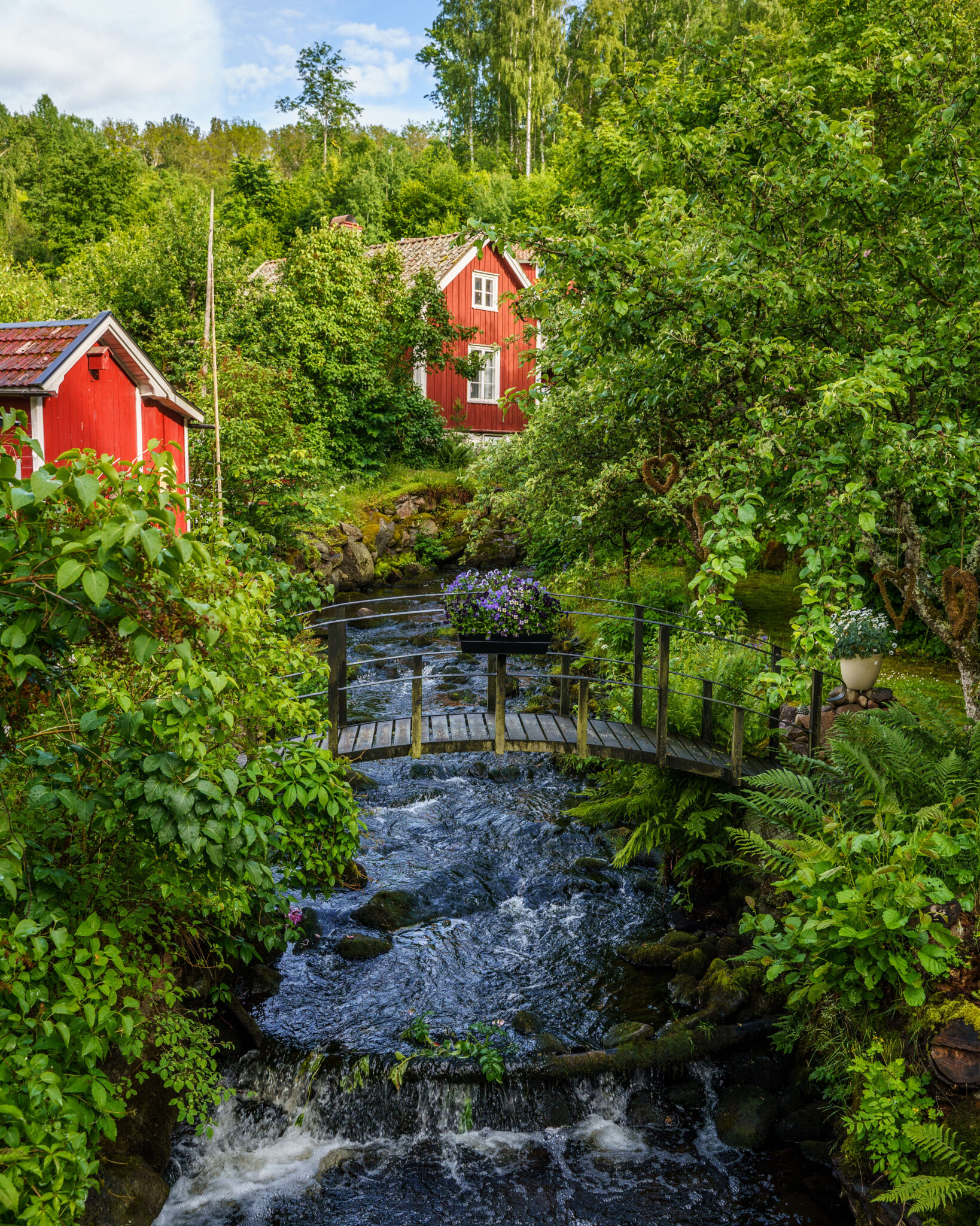 Porlande bäck med liten träbro omgiven av frodig grönska och röda stugor i Röttle by längs Röttlestigen, Småland
