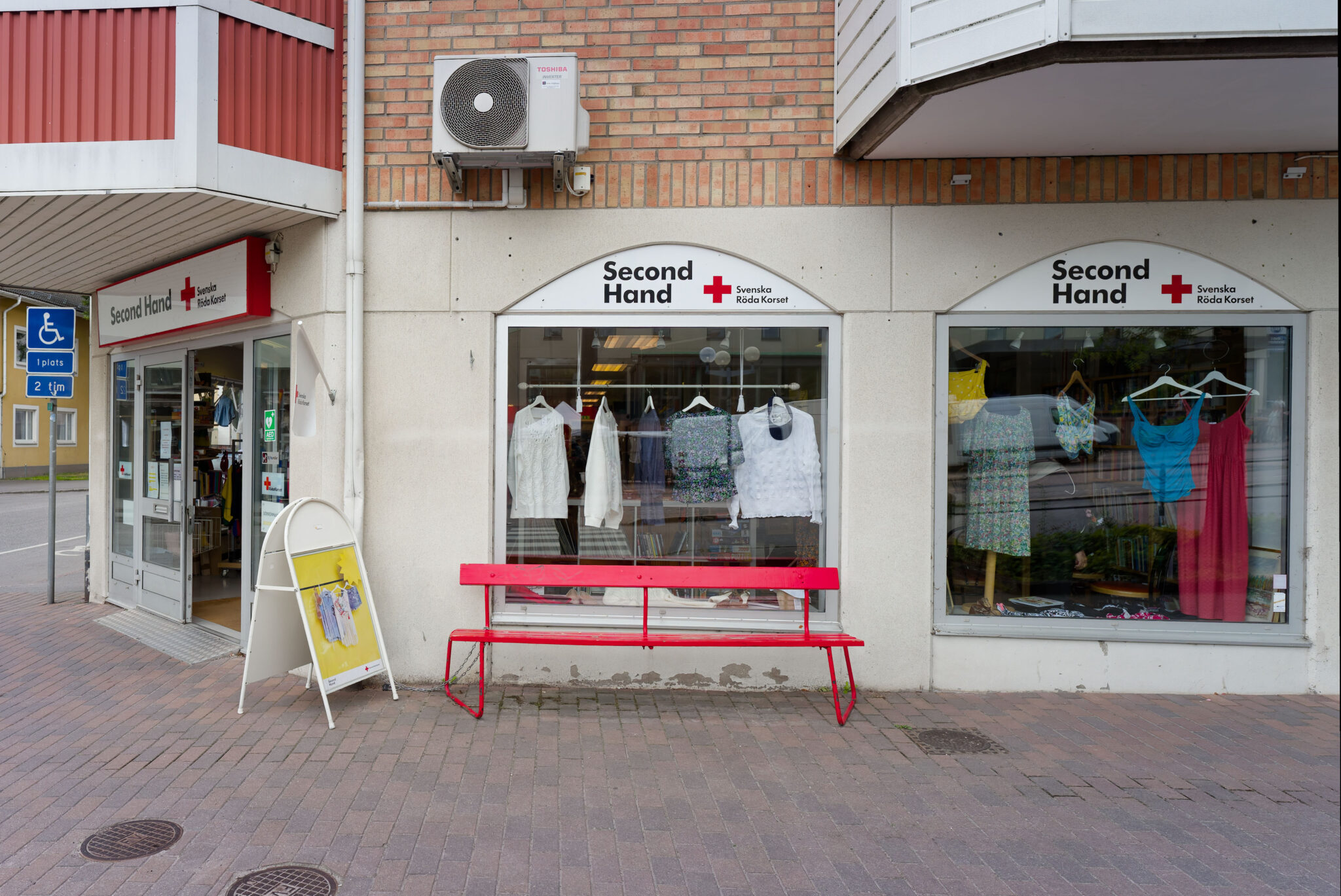 Röda Korset Second Hand-butik i Tranås med röd parkbänk utanför och kläder i skyltfönstret.