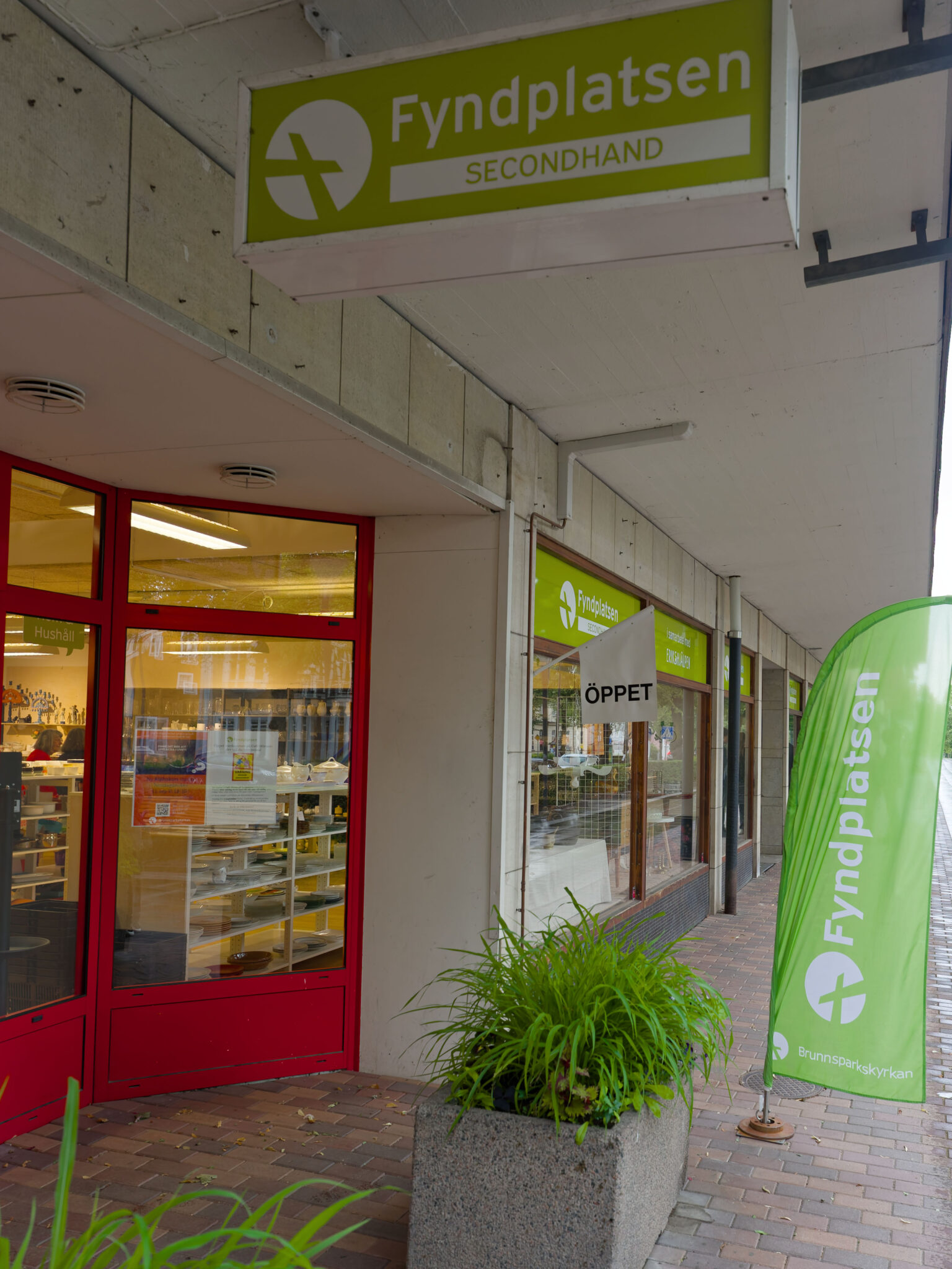 Entrén till Fyndplatsen Secondhand i Tranås med gröna skyltar, röda dörrar och en flagga från Brunnsparkskyrkan.