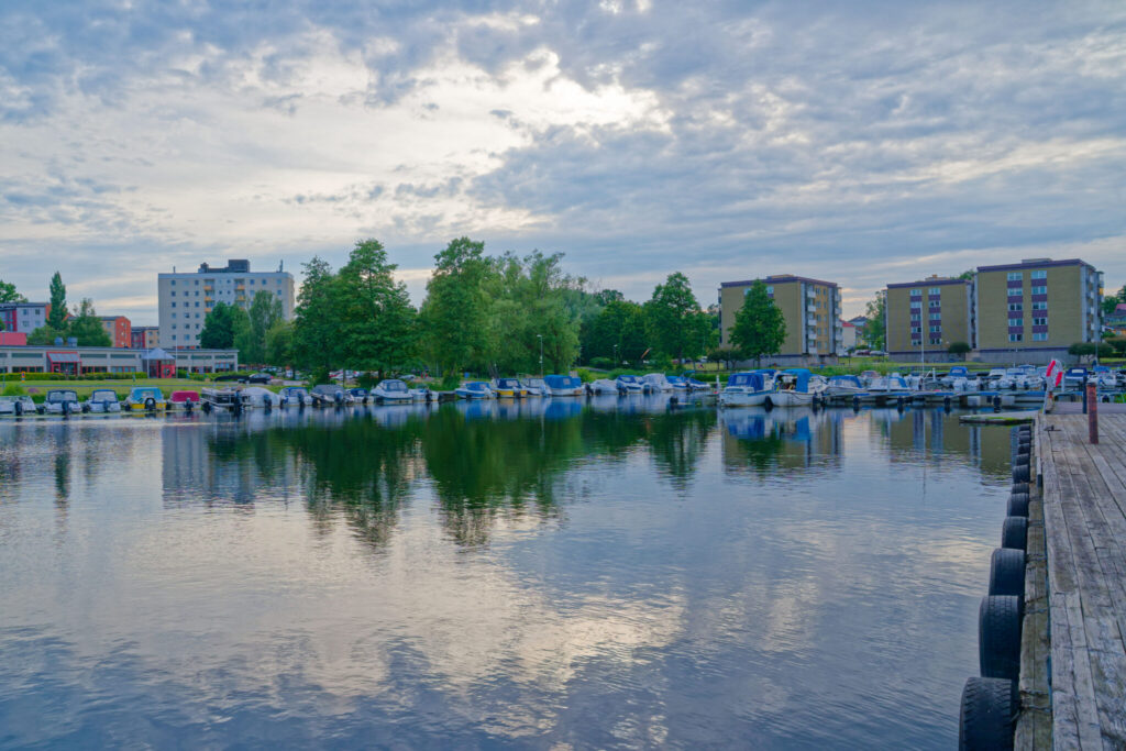 Tranås hamn med båtar förtöjda vid bryggan, spegelblank vattenyta och flerbostadshus omgivna av gröna träd i bakgrunden.