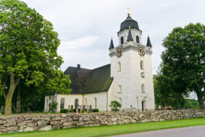 Säby kyrka, en vitputsad kyrka med klocktorn, mörkt tak och en stenmur längs grön kyrkogård.
