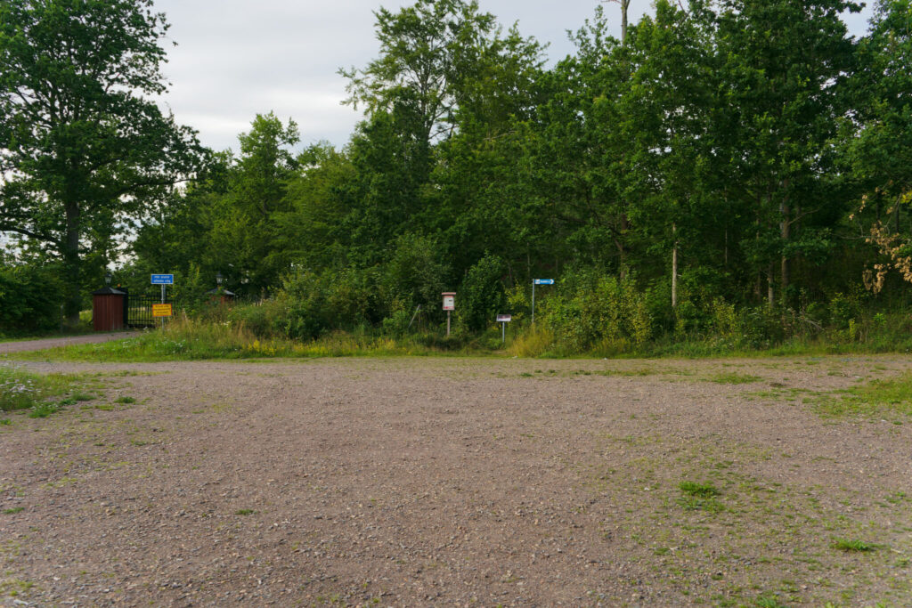 Grusad parkeringsplats vid stigen mot Brahälla slottsruin i Tranås, med informationsskyltar och tät grönska i bakgrunden.