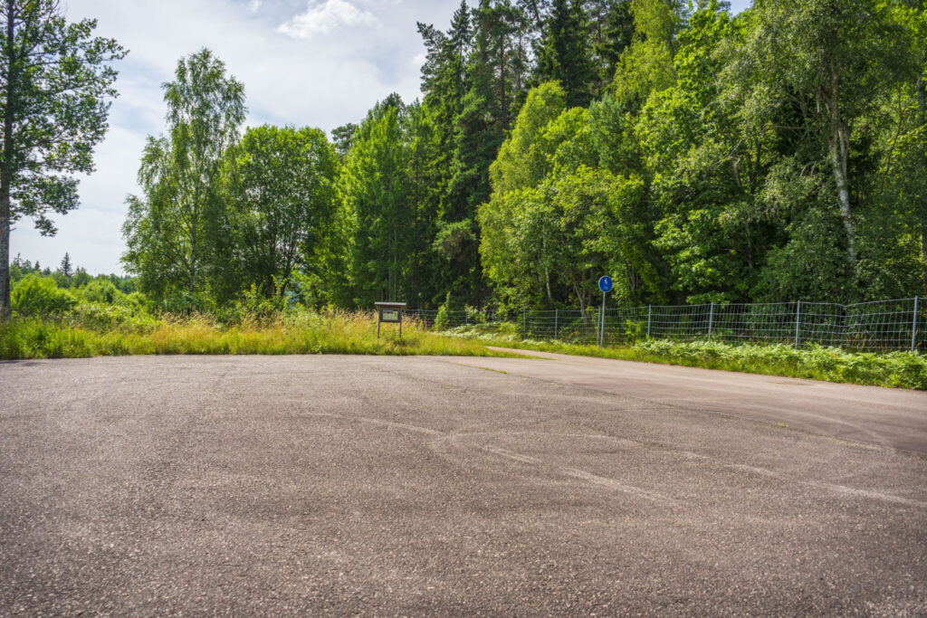 Parkeringsplats vid Holavedens urskog i Tranås kommun, omgiven av frodig sommarskog med björkar och granar.