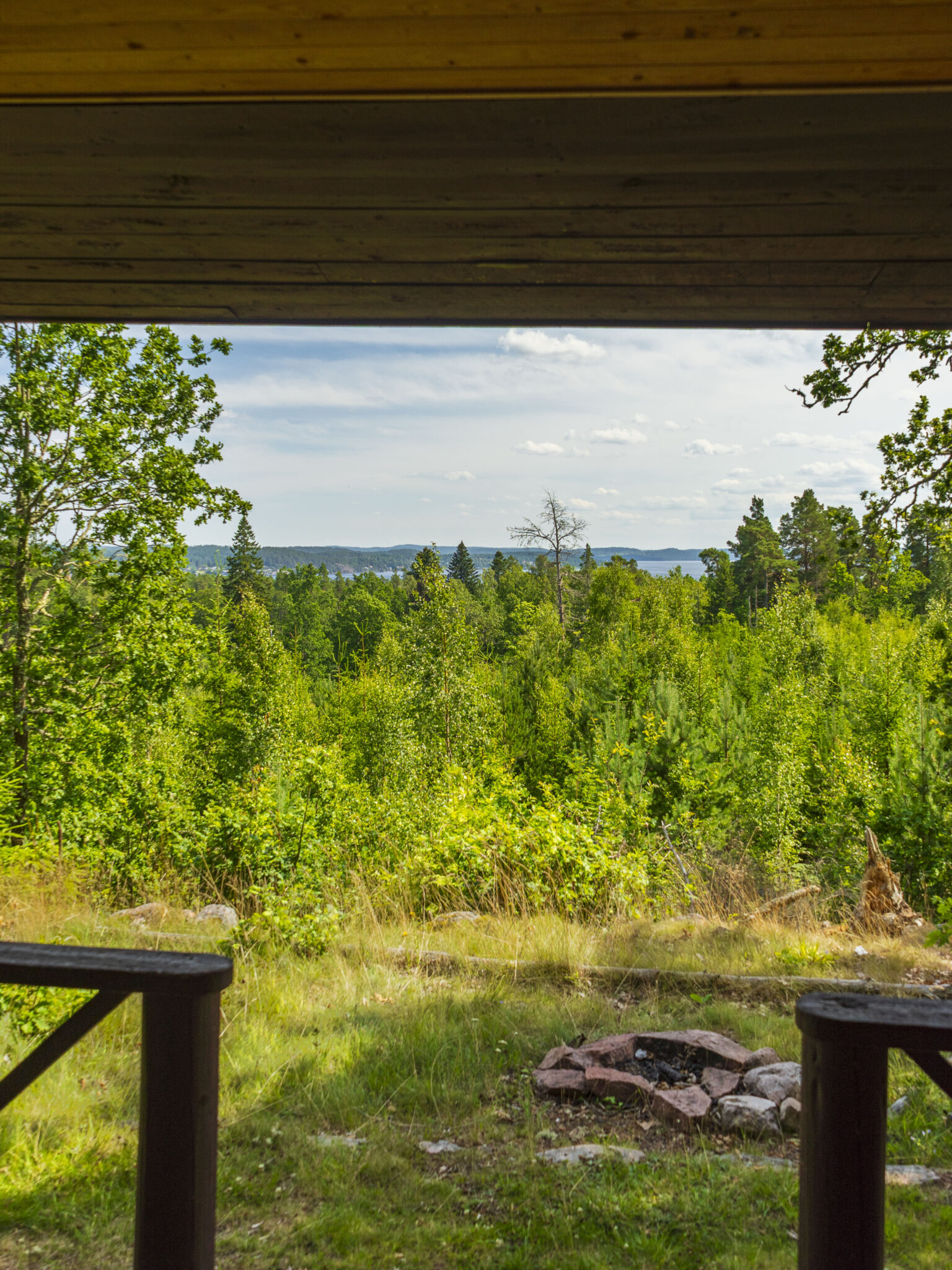 Utsikt från vindskyddad rastplats vid Berners utsikt på Gubbebacken, med eldstad, grön skog och sjöglimt i bakgrunden.