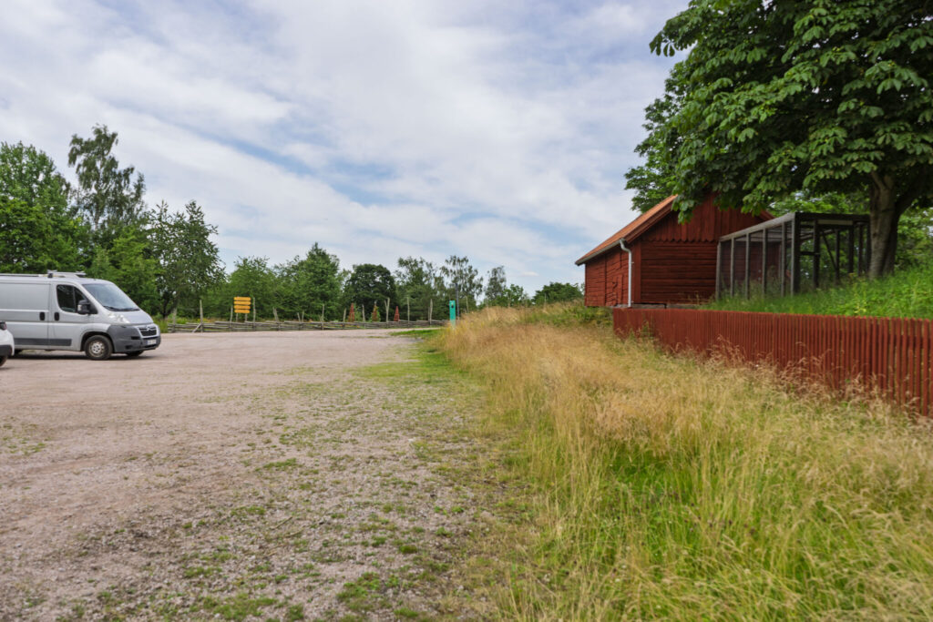 Grusparkeringsplats med vit skåpbil vid röd träbyggnad och högt gräs, omgiven av lövträd i Tranås kommun.